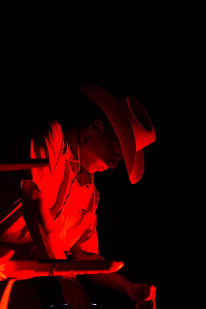 Chuquimamani-Condori performing. Their face tilts downwards. They are wearing a wide brimmed hat, the background is dark and their face is lit dramatically by the cast of a red light