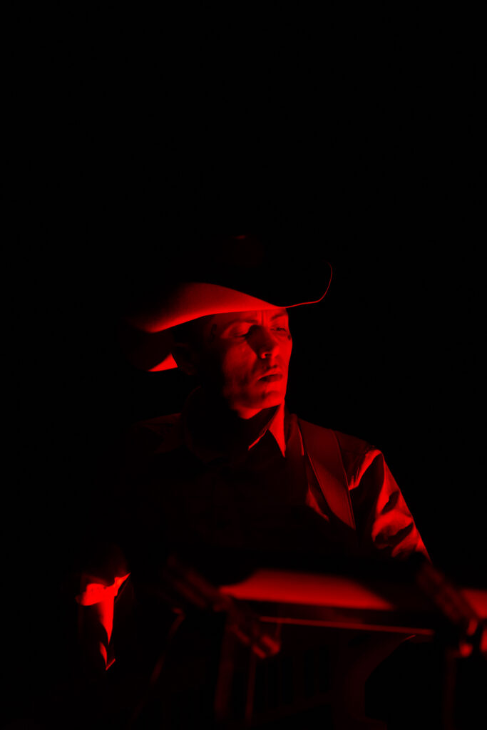 Chuquimamani-Condori performing. They are wearing wide brimmed hat, the background is dark and their face is lit by the cast of a red light