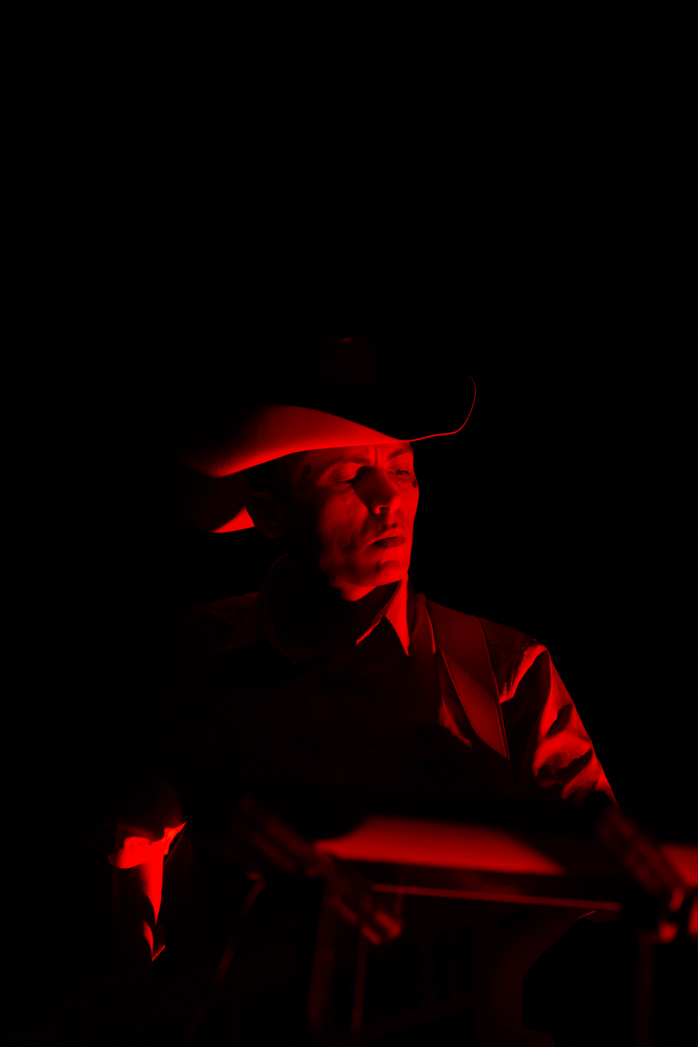 Chuquimamani-Condori performing. They are wearing wide brimmed hat, the background is dark and their face is lit by the cast of a red light
