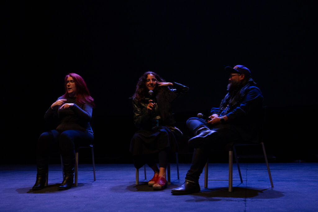 Beatriz Santiago Muñoz on stage in conversation with Pablo de Ocampo. Sat next to them is a BSL interpreter. The background is dark with a cast blue light,