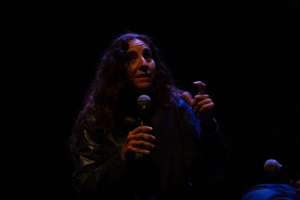 Beatriz Santiago Muñoz speaks into a microphone. The background is dark and their face and hands are dramatically lit.