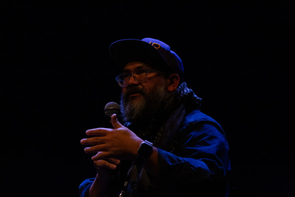 Pablo de Ocampo speaks into a microphone whilst gesturing with their hand. The background is dark and dimly lit, their features are highlighted by the cast of a blue light. The have a beard and wear glasses, a cap tilted upwards and a scarf