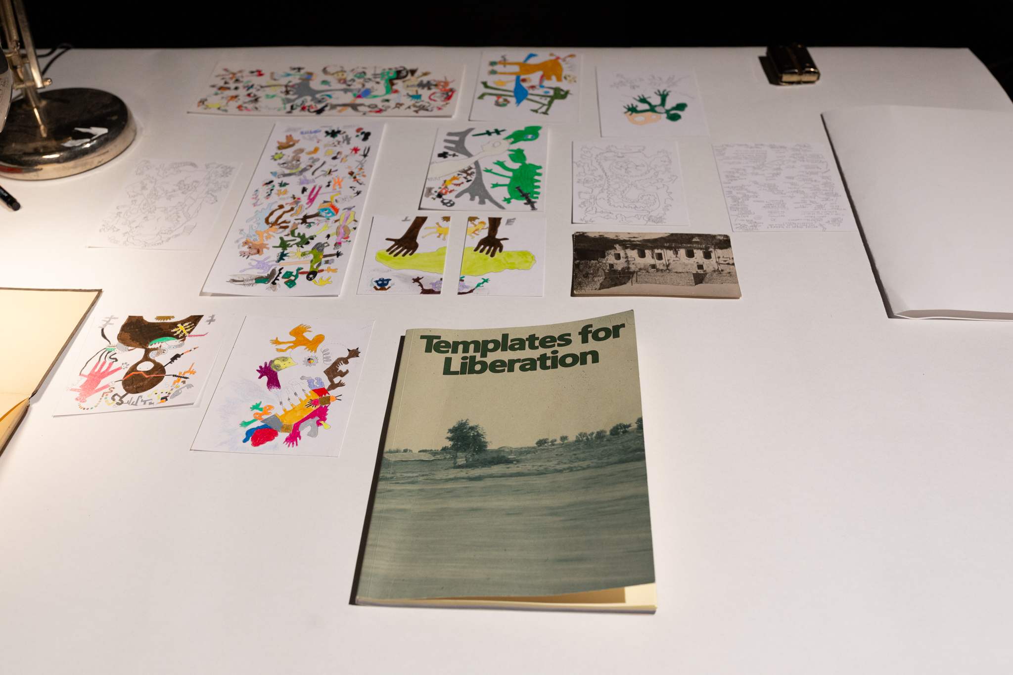 a white table with items laid out on it including book titled "Templates for Liberation", several had pieces of paper with hand drawn colourful illustrations of imagined creatures, hands, writings and an black & white postcard