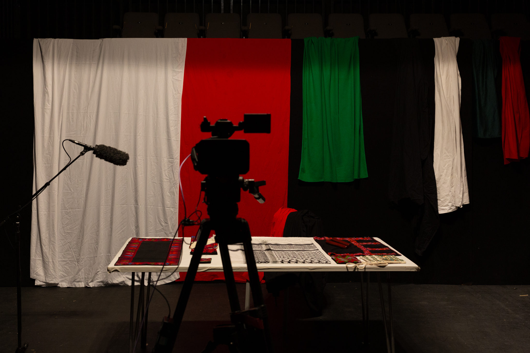 silhouette of a video camera, in the background is green, white & red fabric draped over a screen, in front is a table with carefully folded & placed red & white fabric.