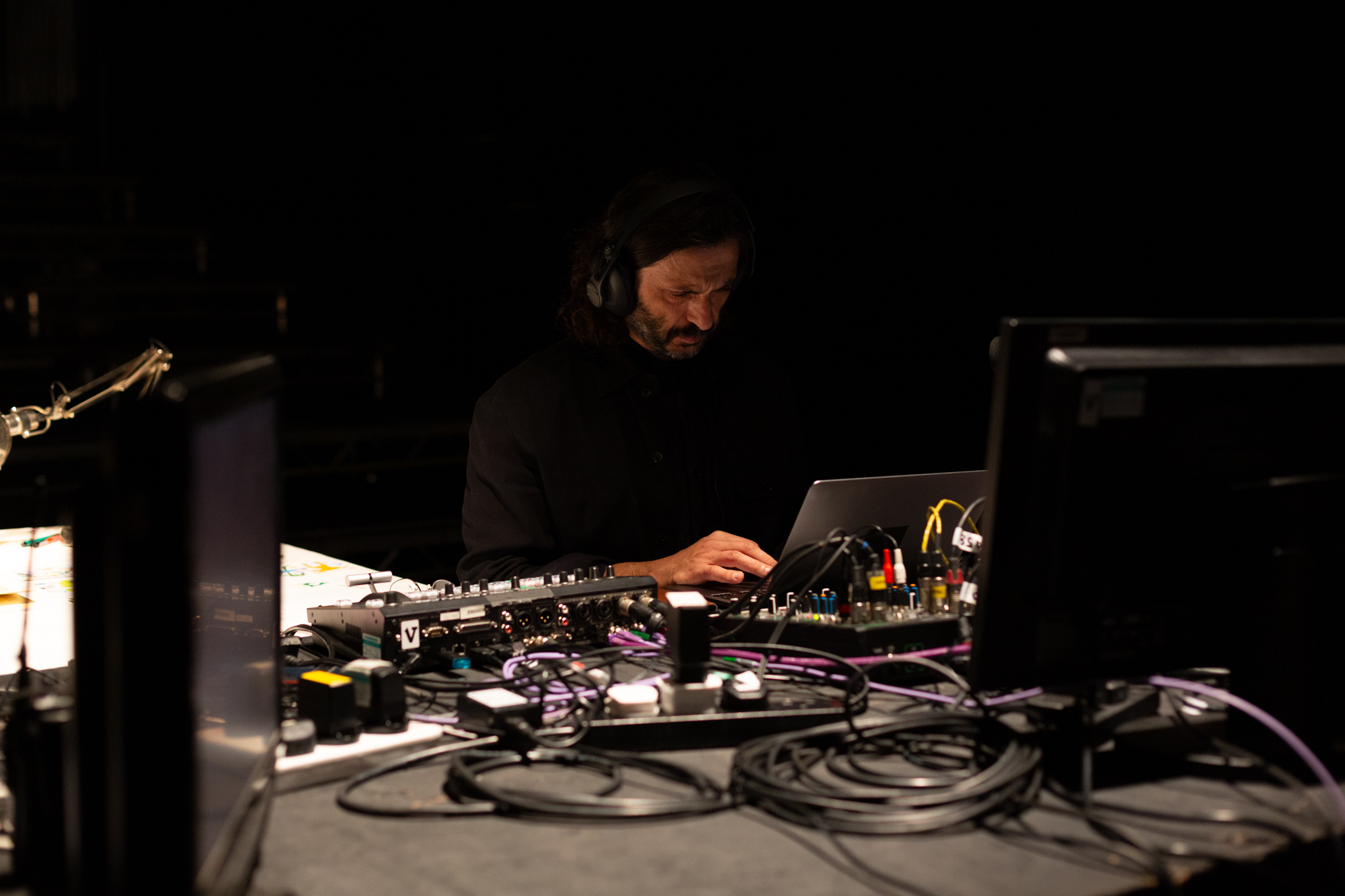 Someone sat at a large desk the are wearing headphone and looking at a laptop. On the desk are many wires, cables & extension leads and a monitor
