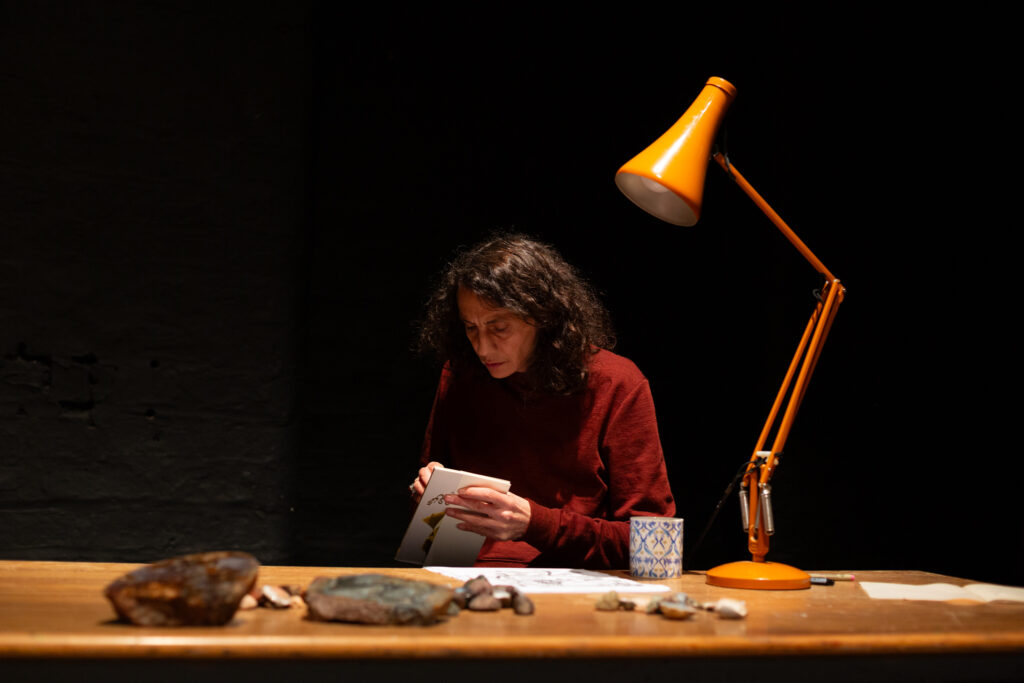Someone sits at a desk writing in a notebook. On the table is yellow poised desk lamp.