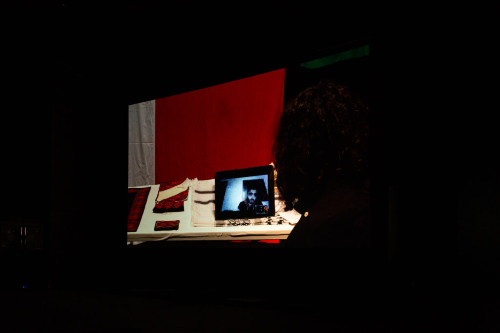 Projected image of a tablet screen featuring someone on a video call. The tablet is sat on a white desk with red and white fabric. To the left we see the back of someone's head as they look at the tablet screen.