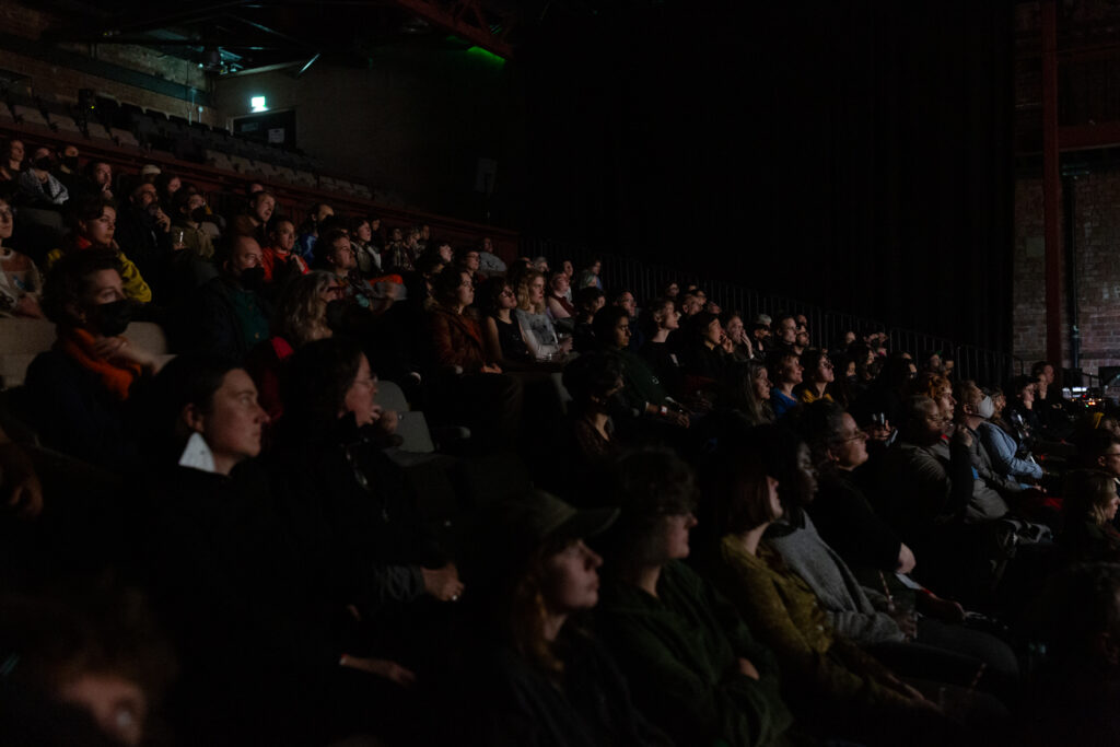 An audience in a seating bank facing forward watching. Their faces highlighted by the glow from a screen.