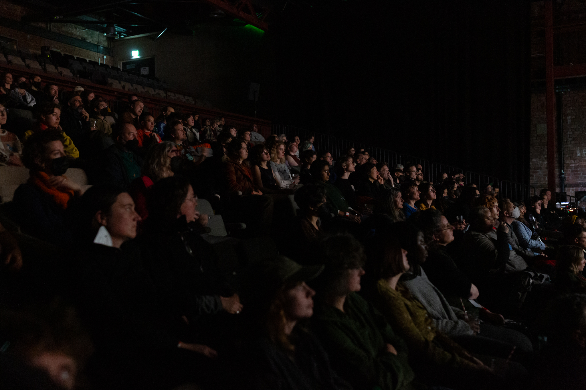 An audience in a seating bank facing forward watching. Their faces highlighted by the glow from a screen.