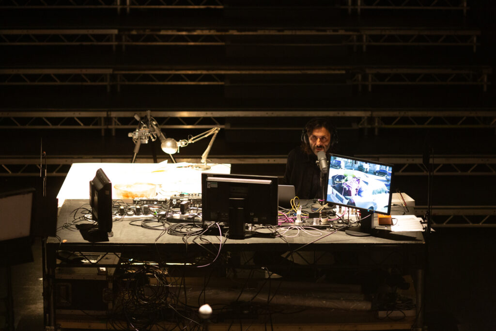 Someone sat at a large desk talking into a microphone. There are several monitors, many cables and extension plugs, a poised desk lamp and video camera