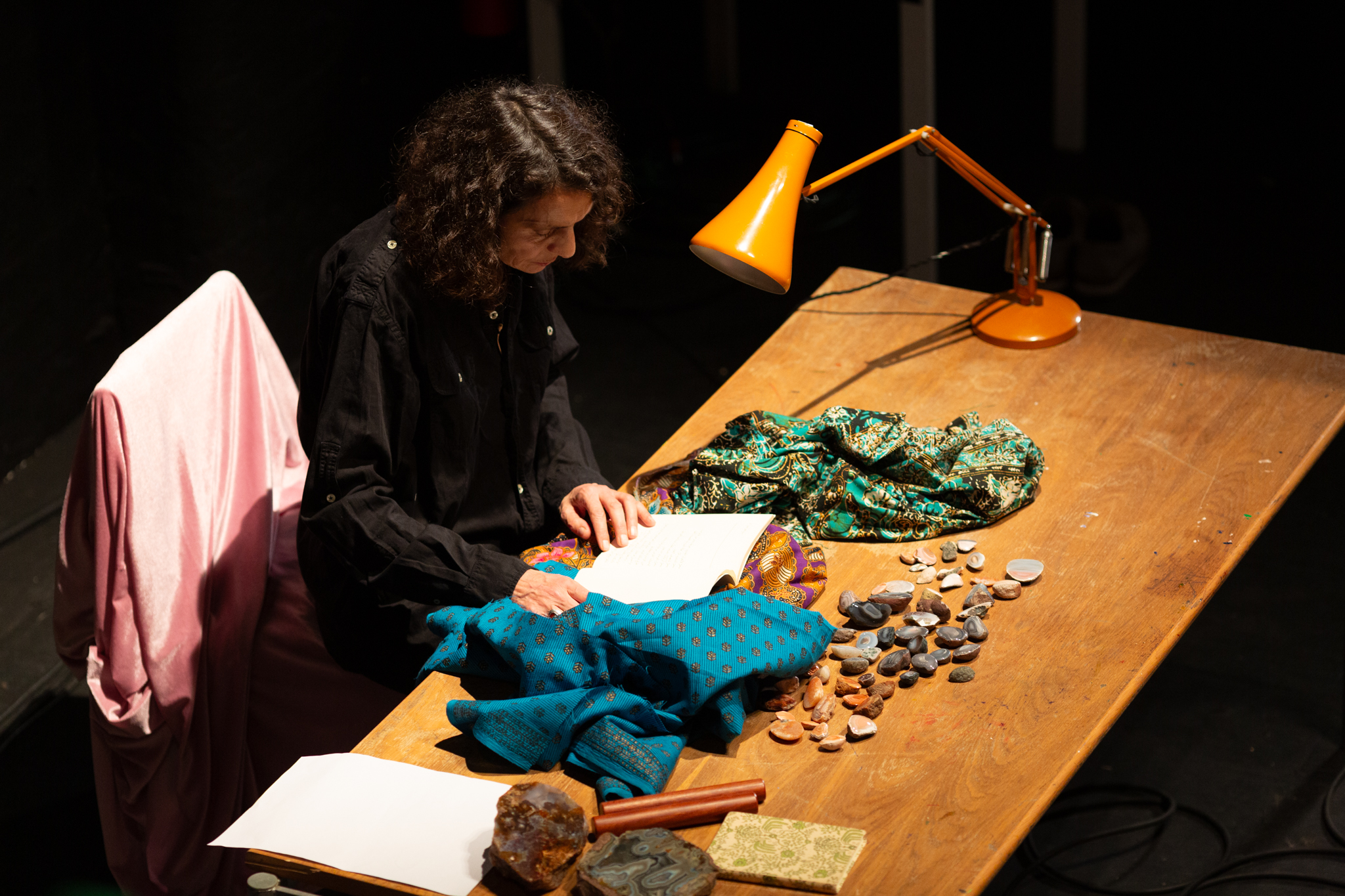Someone sat at a desk with a book open in front of them and a yellow desk lamp. The desk is strewn with items like patterned fabric and polished stones