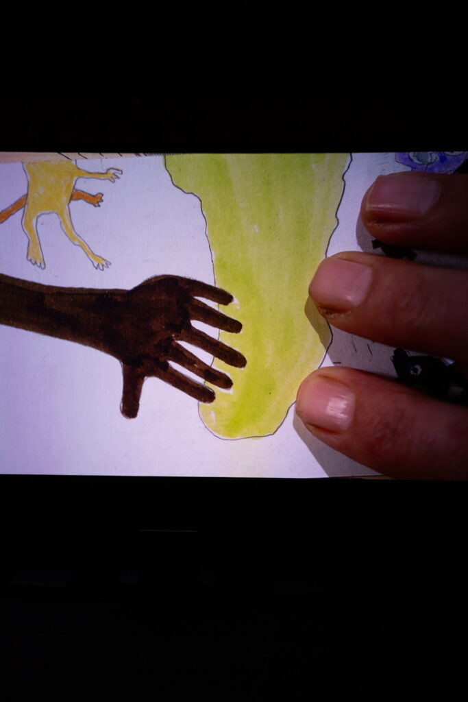 Projected image of fingers sat on top of a child-like drawing of a hand with six fingers on top of a green shape that looks like a map