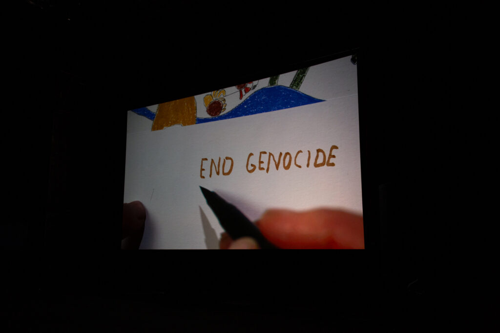 A projected screen, in the image a hand writes 'end genocide'