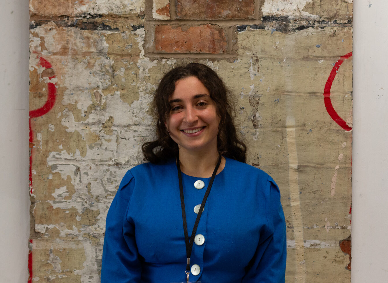 Masa Nazzal smiles, waring a bright blue jumper with oversized pearl buttons. They standi infront of a brick wall.