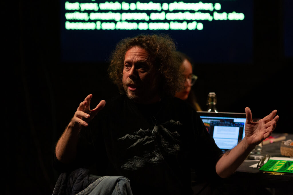 Amilcar Parker talks while gesturing expressively with his hands. He looks towards the audience on his right, and is sat at a table which has a laptop and other items on it.