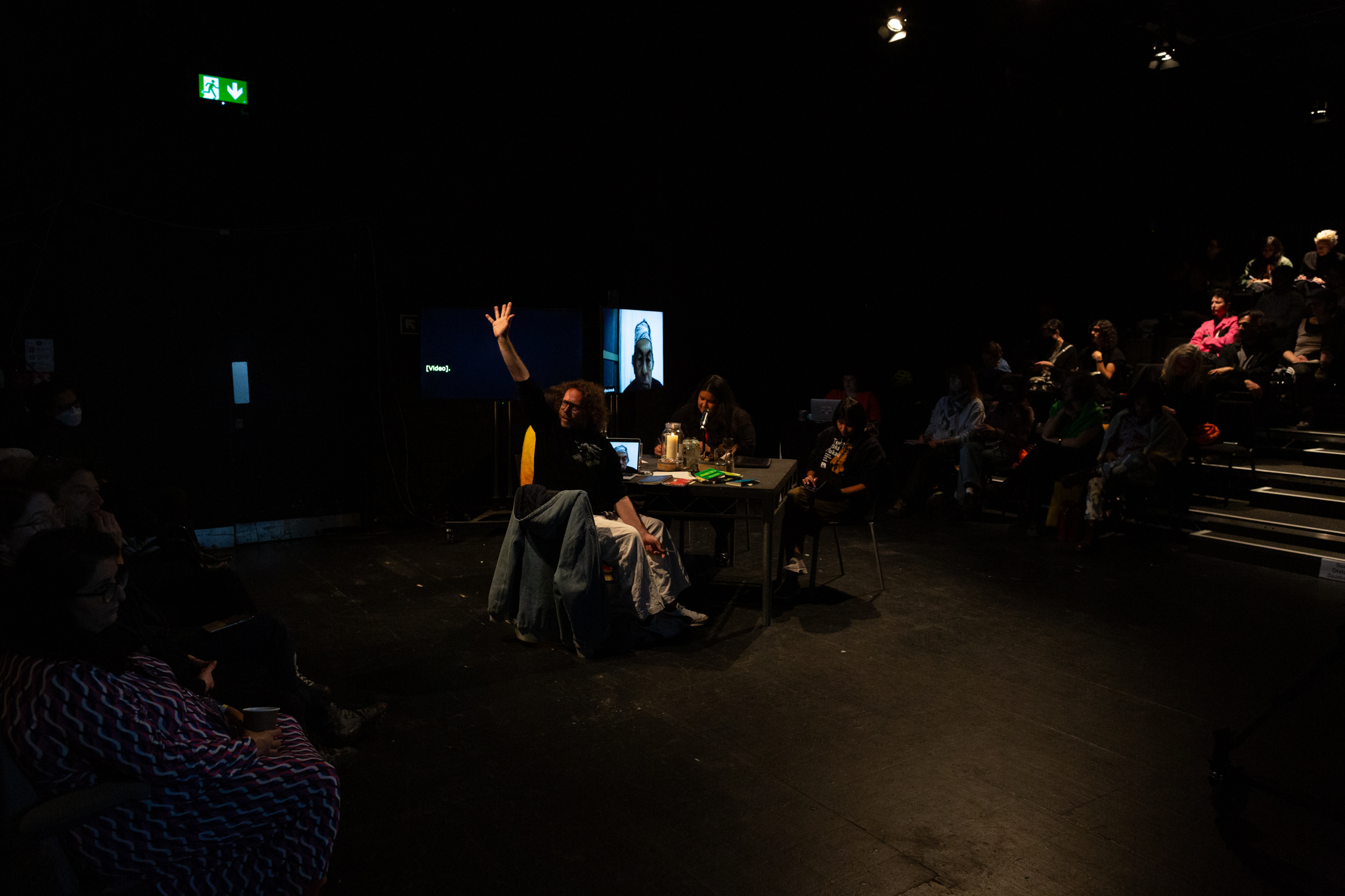 A black box theatre space, in the centre is a black table with four people sat around it. Amilcar Packer expressively holds his up with the palm open facing the audience to his right. Audience are sat in a seating bank to the left & right. of the table
