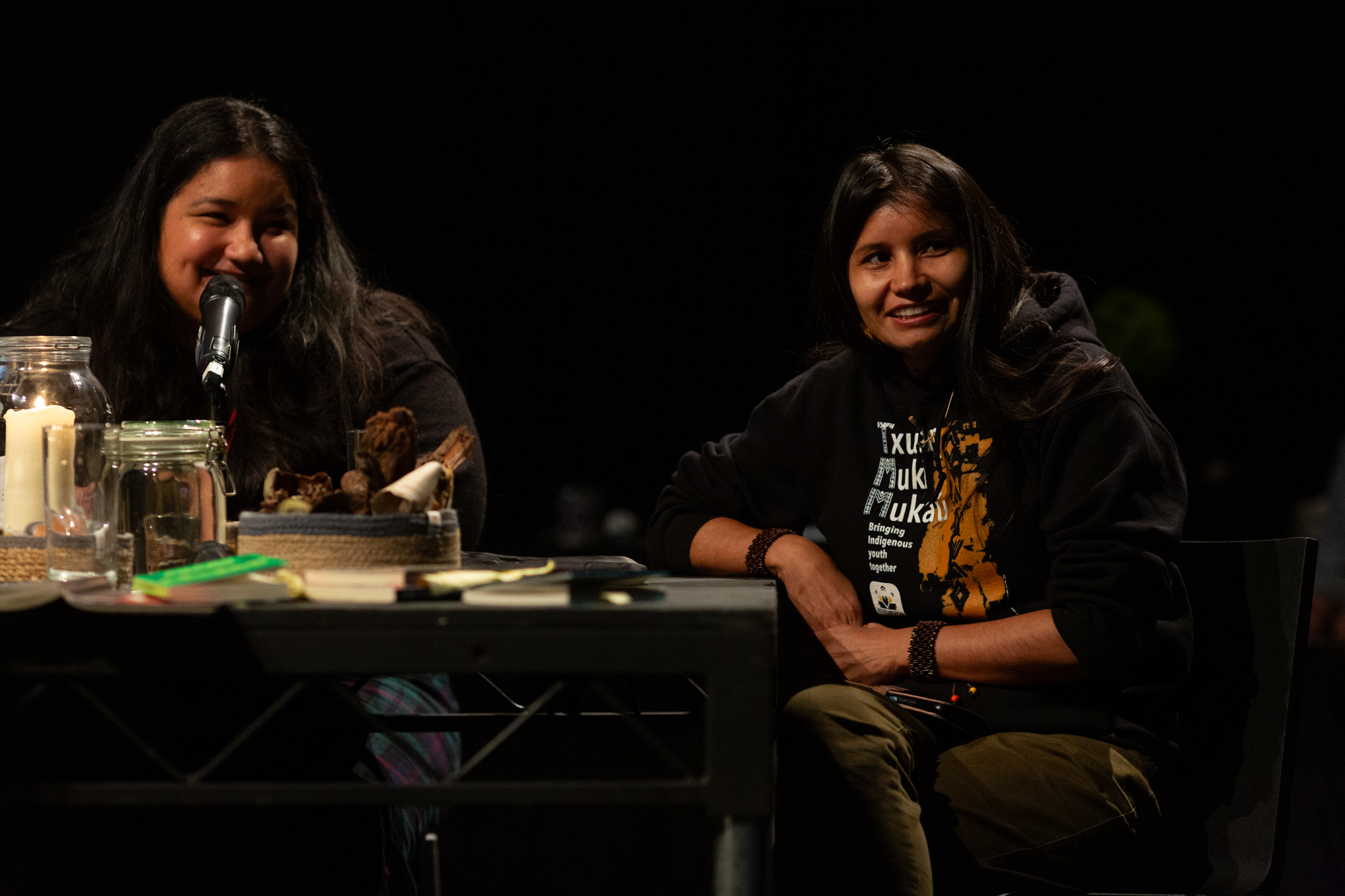Arissana Pataxó sits smilling at a discursive event. She sits at a black table, next to her sits the language interpreter who translated the event from Brazilian Portuguese to English.