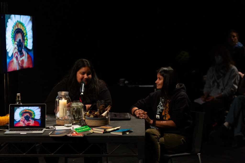 Arissana Pataxó smiles while she sits at a large black table with an audience behind her and a language interpreter next to her. On the table is a laptop with a drawn image of a person wearing an indigenous feather head dress, taking a photo on a camera that points towards the viewer. The image on the laptop is mirrored on a larger screen for the audience to view.