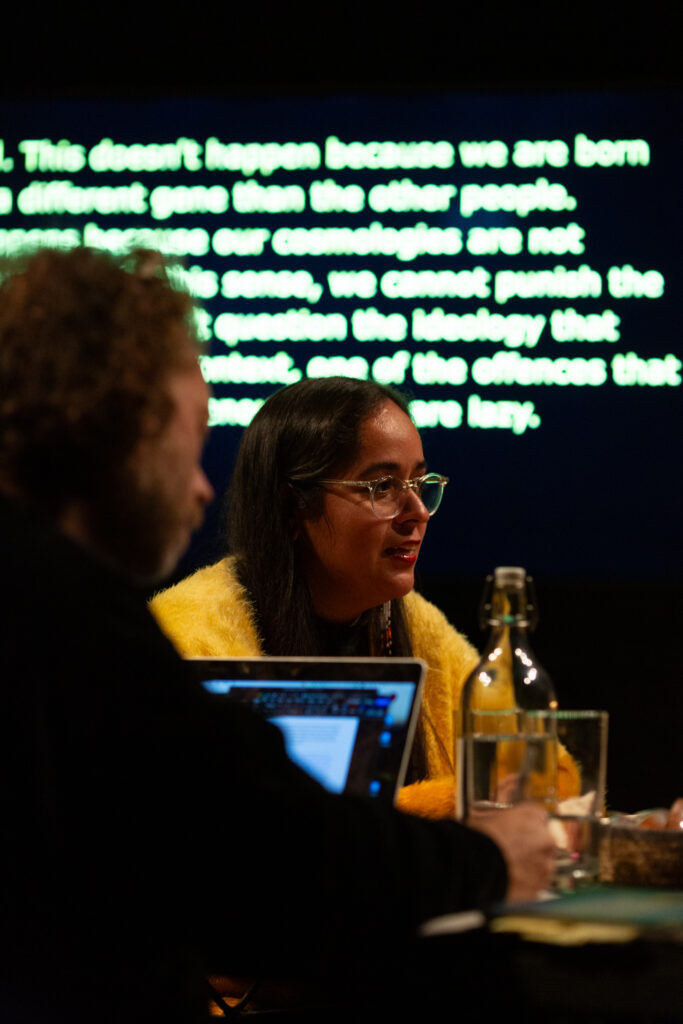 Geni Núñez talks at a discursive event. She wears a yellow cardigan. They are sat at a table which has a glass water bottle and a laptop. Behig them is a screen with blurred greenish white text