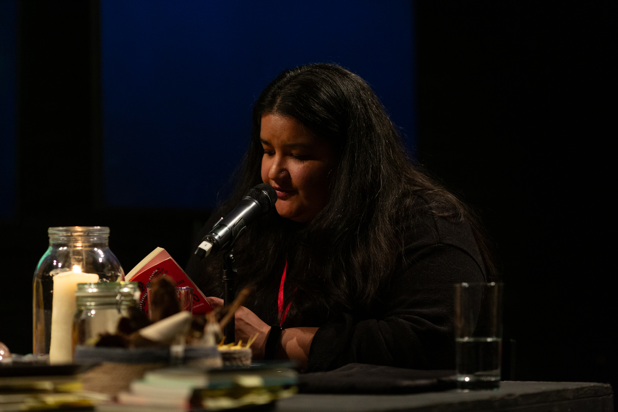 The language interpreter reads a passage from a book. They speak into a microphone that sits on a black table which has candles, glass jars, and drinks on the table