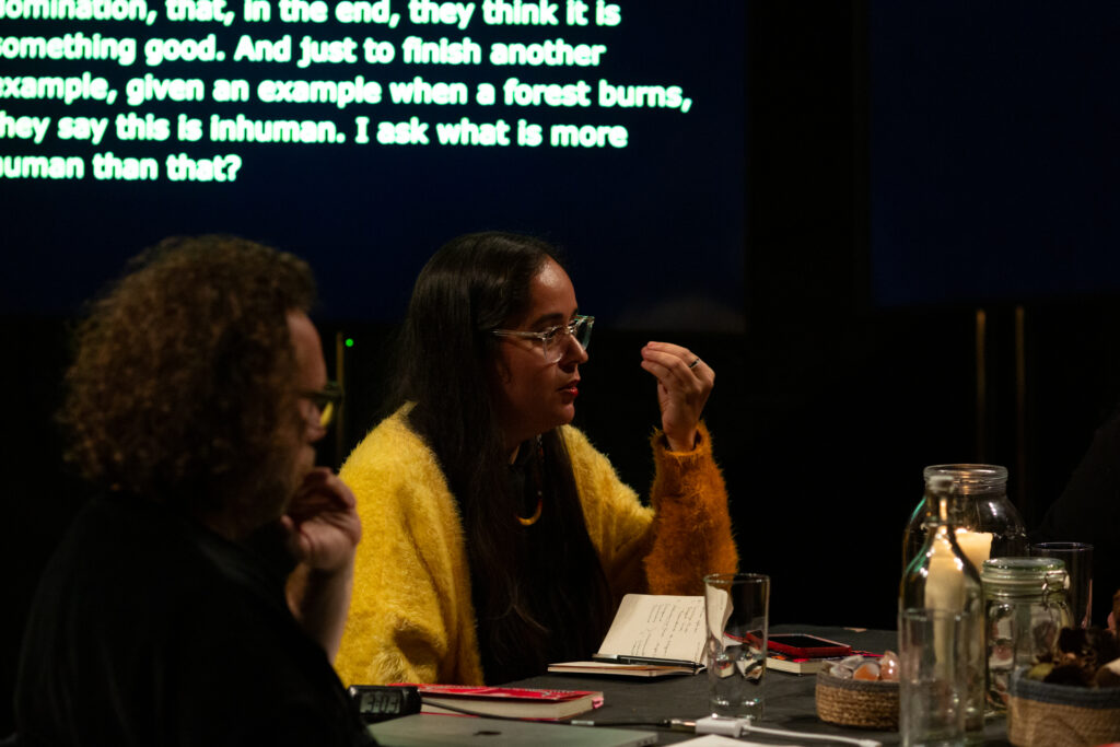 Geni Núñez wears a yellow sweater and gestures thoughtfully with her hand while speaking at a table discussion. She has a notebook open, and a screen behind her displays text about human nature. Candles and jars are on the table.