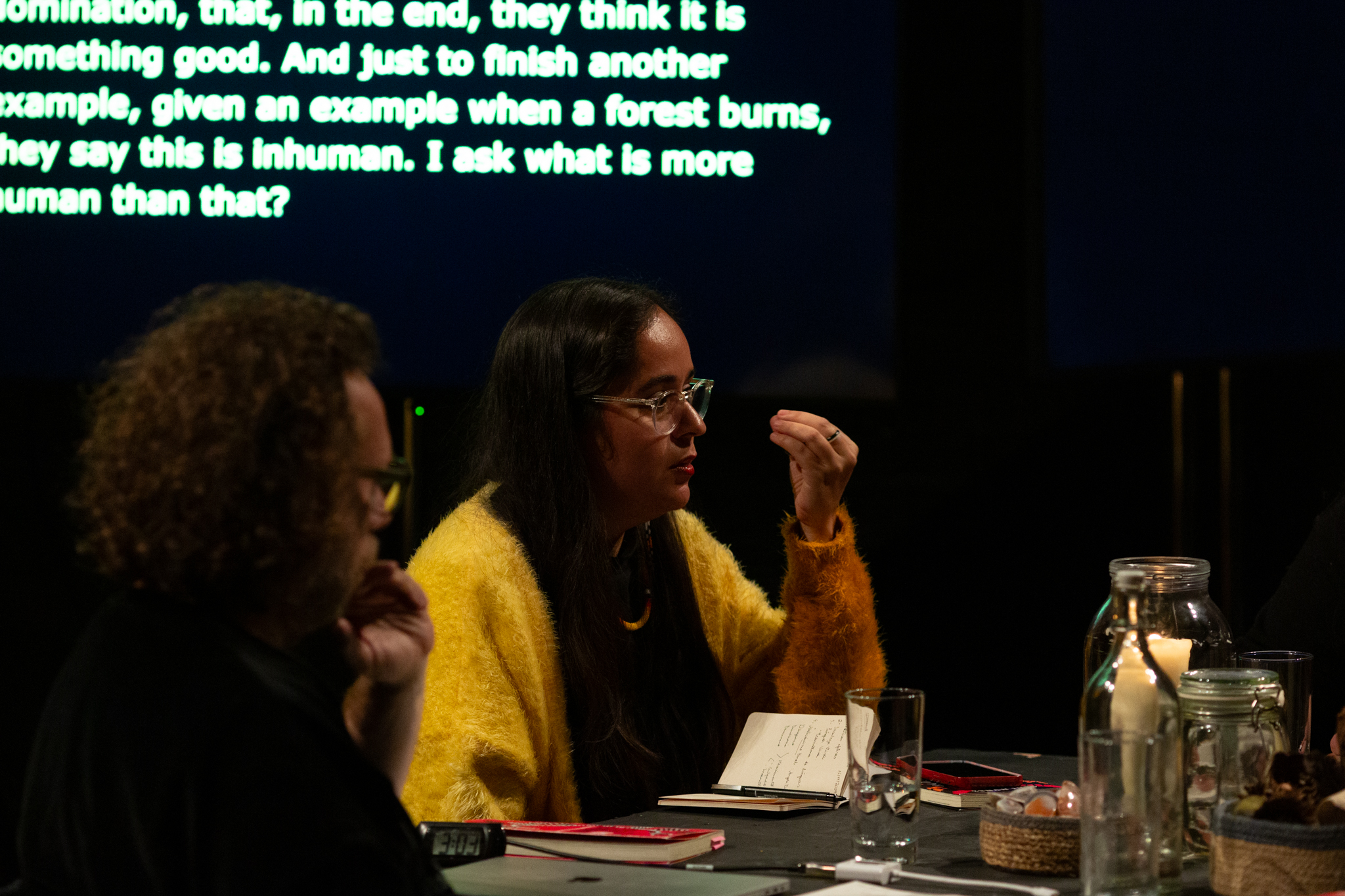 Geni Núñez wears a yellow sweater and gestures thoughtfully with her hand while speaking at a table discussion. She has a notebook open, and a screen behind her displays text about human nature. Candles and jars are on the table.