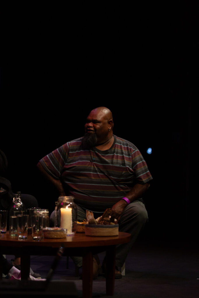 Rex Sing from Karrabing sits on stage in a relaxed posture. The background is dark and in front is a low table with drinks, candles and small basket.