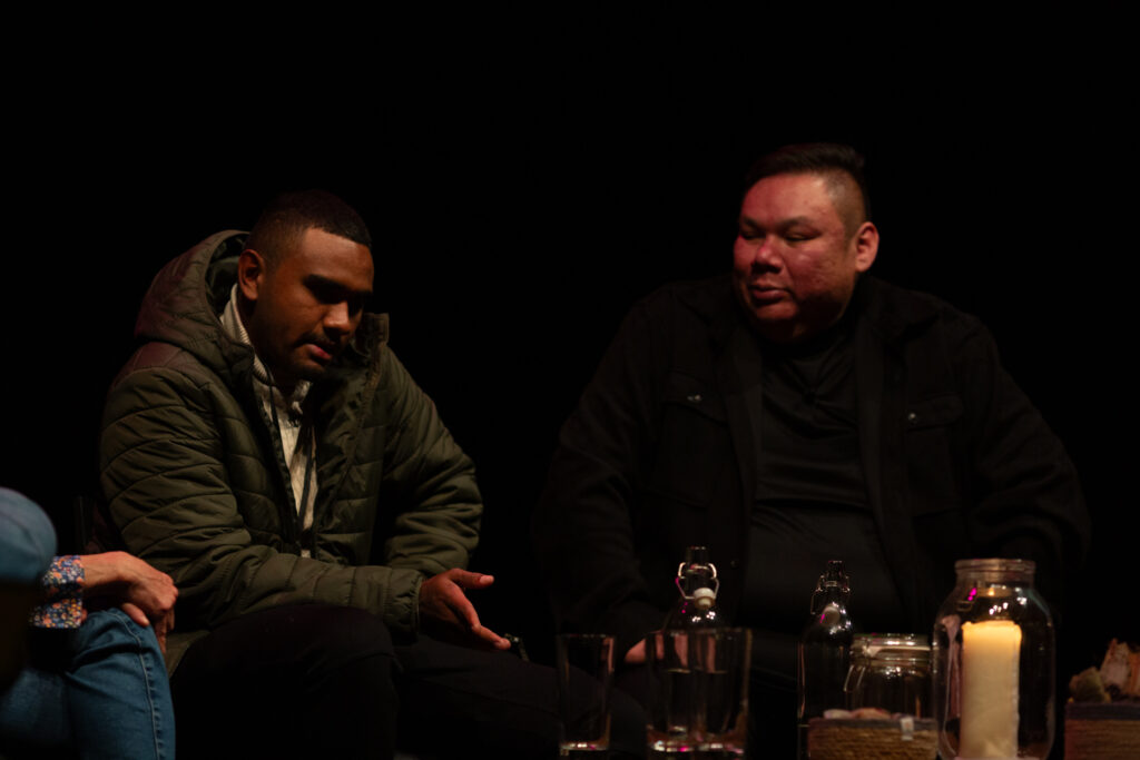 Aiden Sing from Karrabing speaks on stage with Elwood Jimmy. The background is dark and dimly lit.
