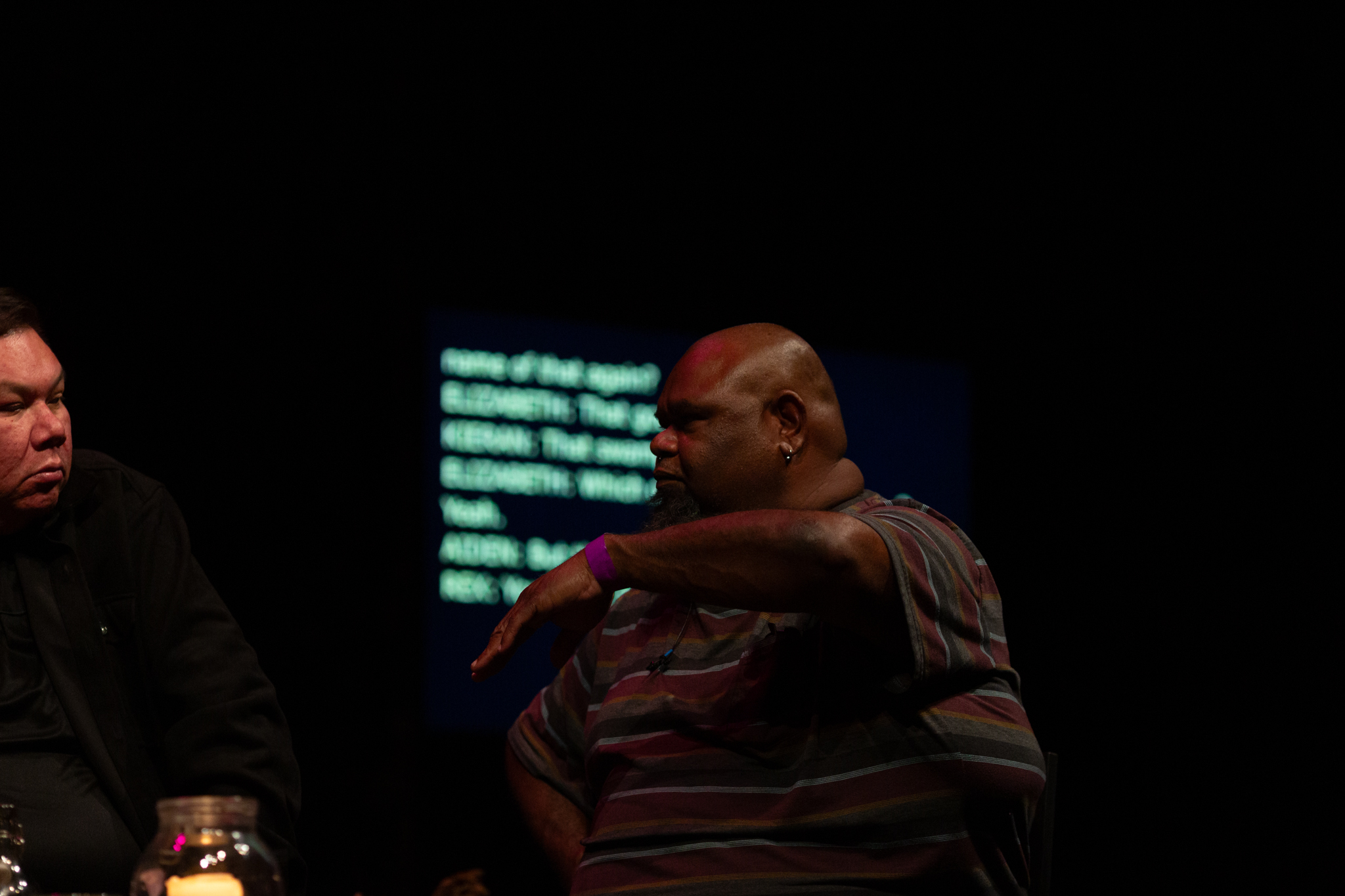 Rex Sing sits one stage expressively gesturing with his hand out in front of him, bent a a right angle at elbow with the palm of the hand facing down at an angle.