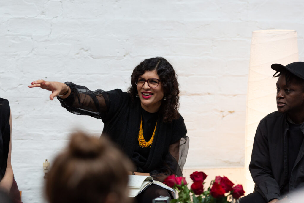 Nat Raha points an arm out in front of her, she is smiling. Nat wears a black outfit with a bright chunky beaded yellow necklace and wears bright red lipstick.