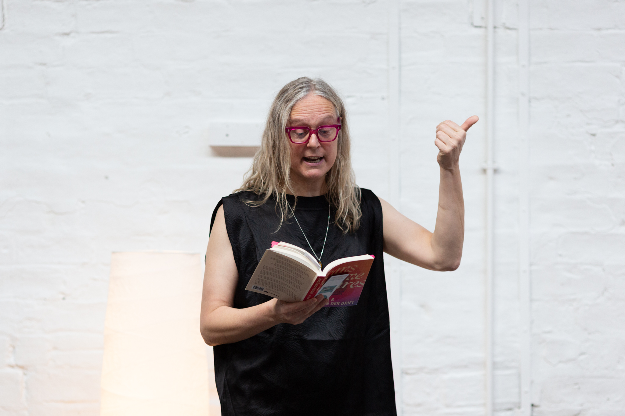 Mijke van der Drift stands reading from a book held in their hand. Mijke wears bright pink rimmed glasses and stands expressively holding an arm out with their thumb in the air