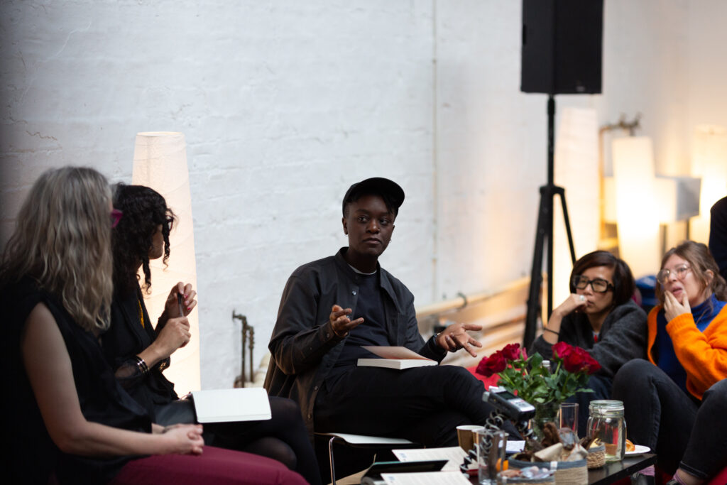 Jay Bernard in a discussion event with Nat Raha and Mijke van der Drift. Jay looks at Nat & Mijke with hand outwards and open in an expressive manner. To the left of Jay are audience members sat listening pensively.