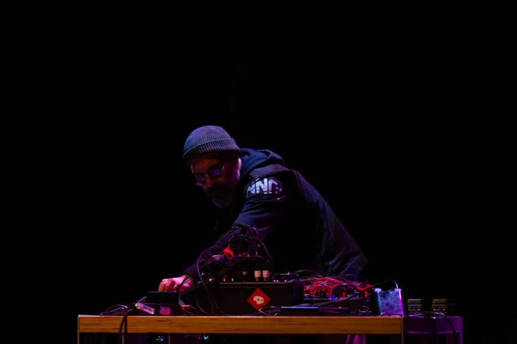 Rashad Becker on stage, performing at a desk with sound and audio equipment. The background is dark and a pinkish red light highlights Rashad