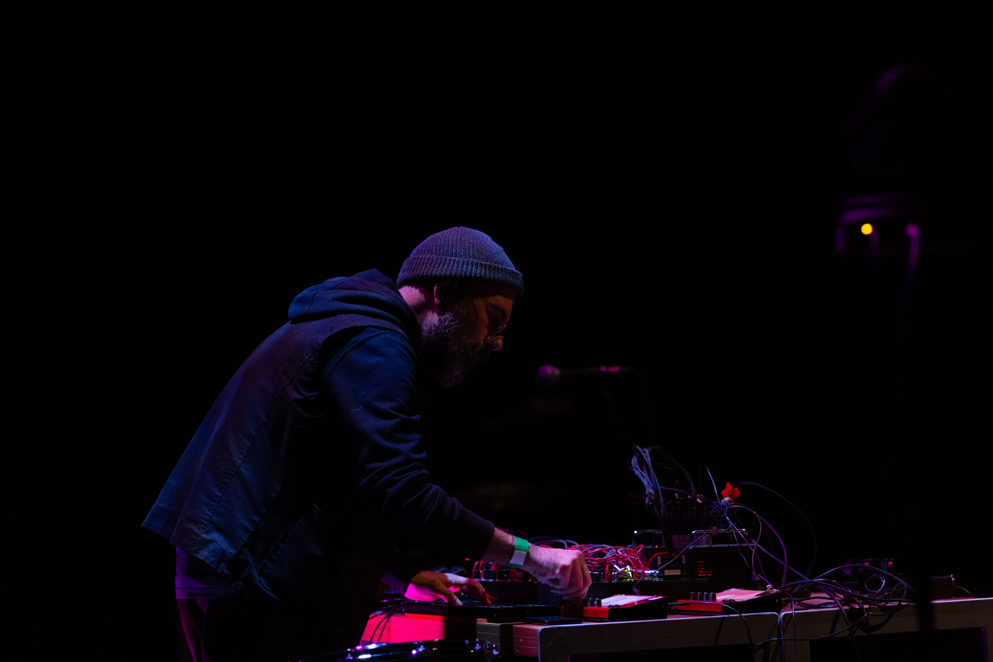 Rashad Becker on stage, performing at a desk with sound and audio equipment. One hand is turning knobs on the sound equipment. The background is dark and a pinkish red light highlights Rashad