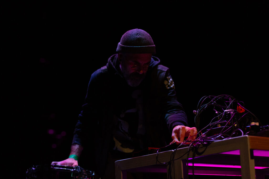 Rashad Becker on stage, performing at a desk with sound and audio equipment. The background is dark and a pinkish red light highlights Rashad