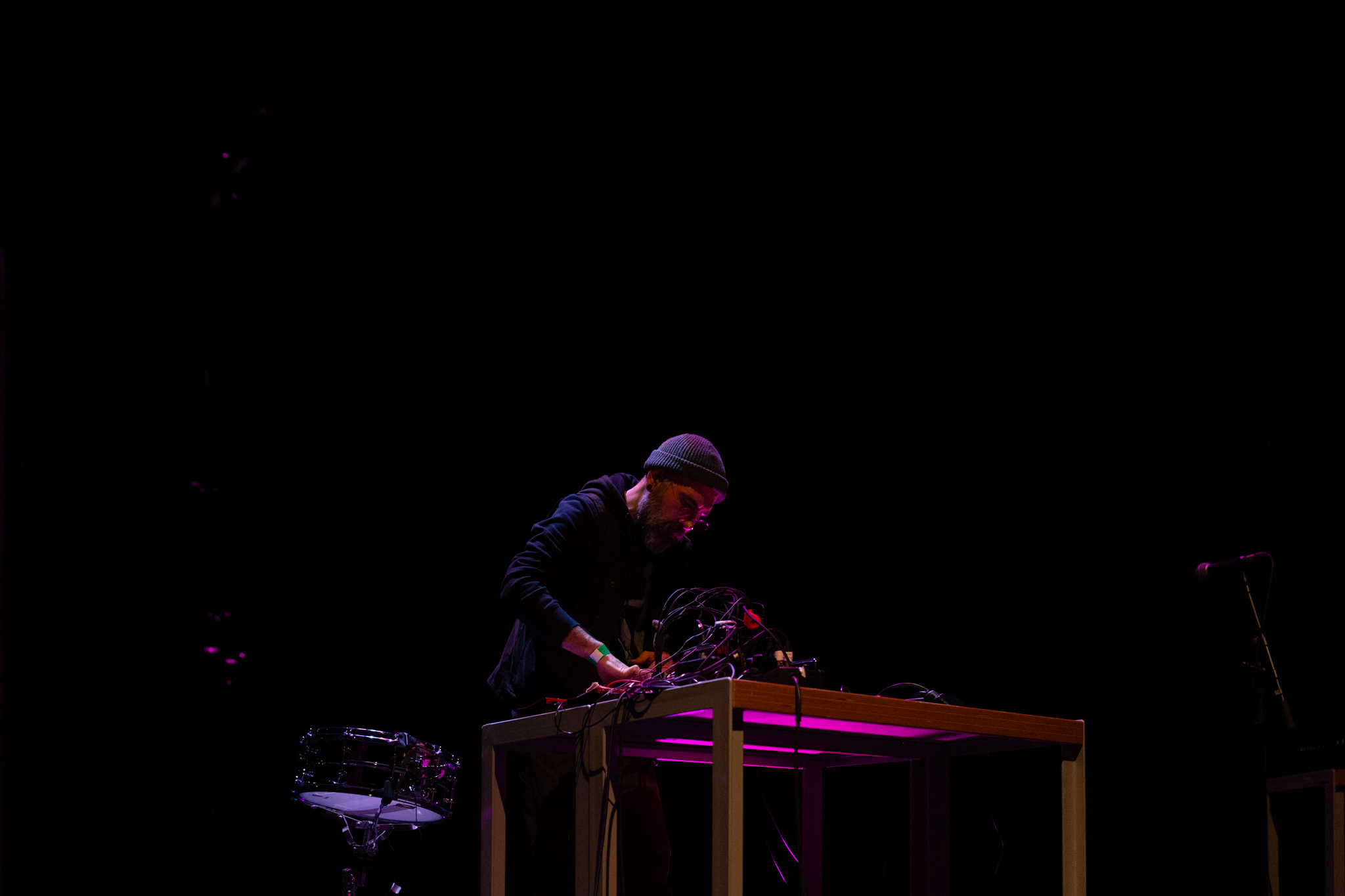 Rashad Becker on stage, performing at a desk with sound and audio equipment. The background is dark and a pinkish red light highlights Rashad