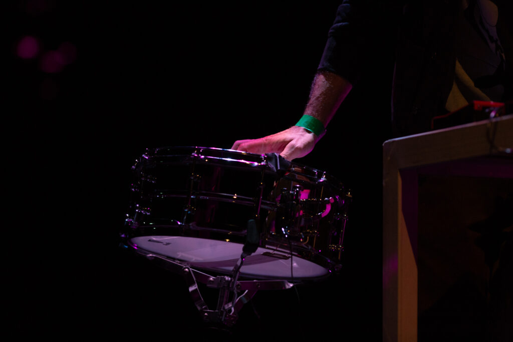 A hand plays a drum. The person is wearing a green wrist band. The background is dark and the hand is dramatically lit in comparison