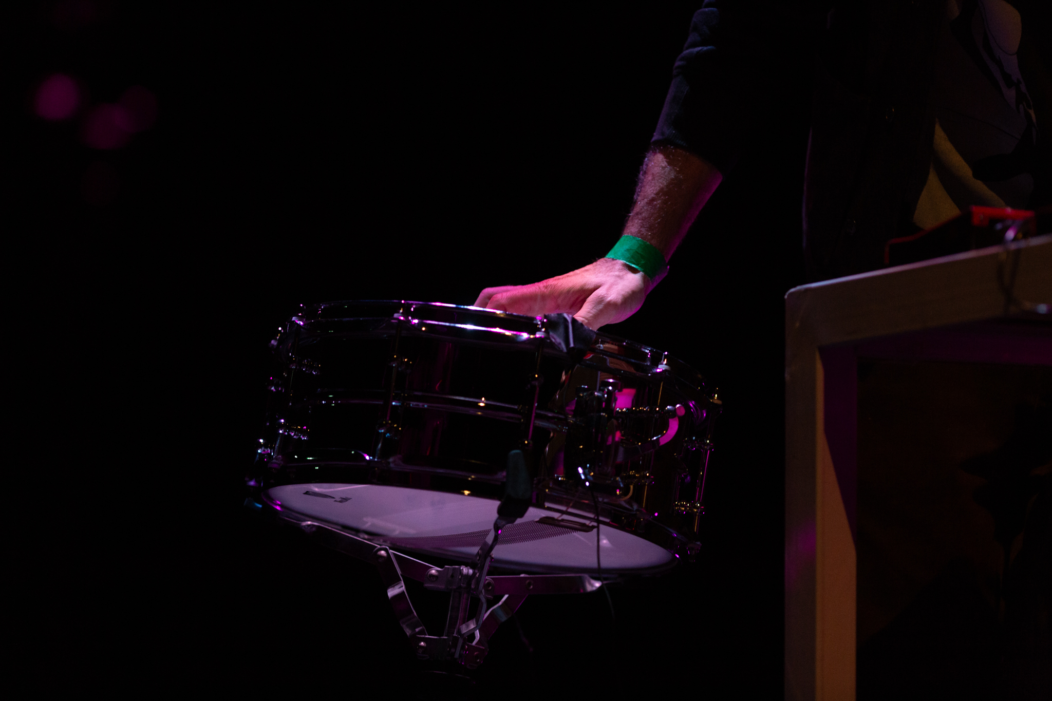 A hand plays a drum. The person is wearing a green wrist band. The background is dark and the hand is dramatically lit in comparison