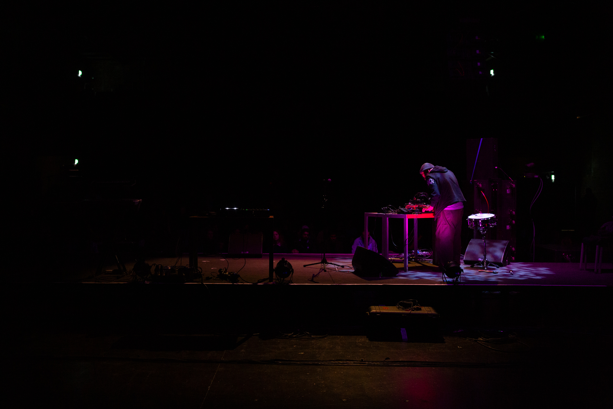 Rashad Becker on stage, standing at a desk with sound and audio equipment. The background is dark and a pinkish red light highlights Rashad