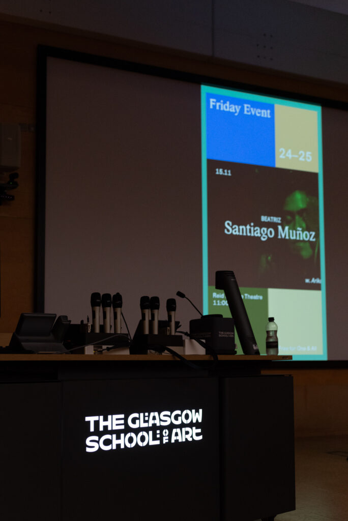 A table with microphones has bright white letters reading "Glasgow School of Art". being the table is a projected screen with an image that says; Friday Event, 24-25, Beatriz Santiago Muñoz