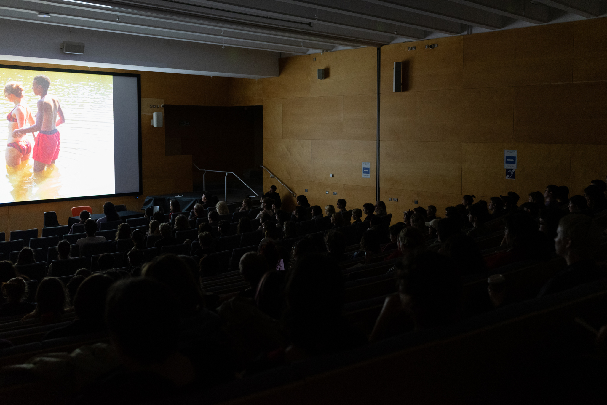A lecture theatre busy with people seated watching a film on a screen. On the screen are two people standing thigh deep in greenish water, perhaps a river.