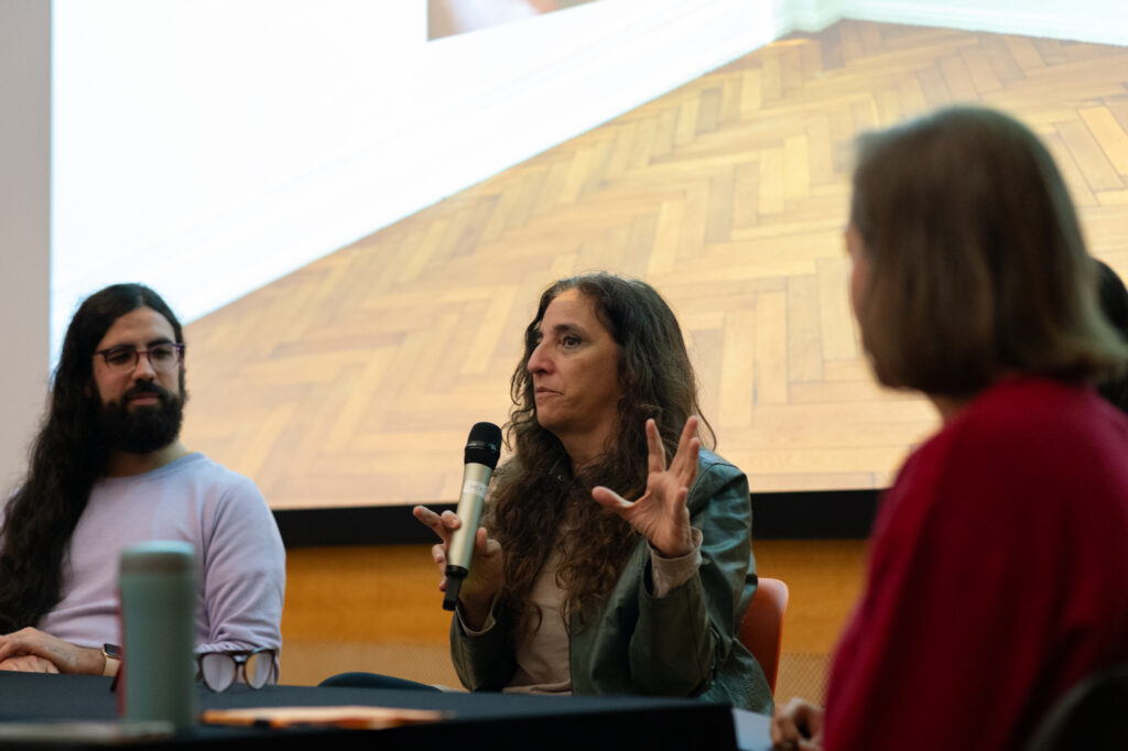 Beatriz Santiago Muñoz talks into a microphone whilst seated in front of an audience. At the back we can see the bottom of a projected image which has a herringbone patterned wooden floor. Three other people are sat next to Beatriz listening intently