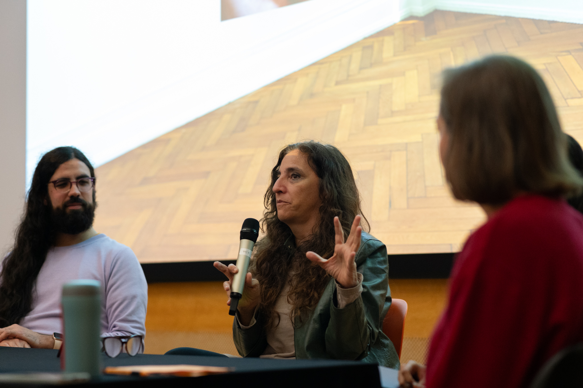 Beatriz Santiago Muñoz talks into a microphone whilst seated in front of an audience. At the back we can see the bottom of a projected image which has a herringbone patterned wooden floor. Three other people are sat next to Beatriz listening intently