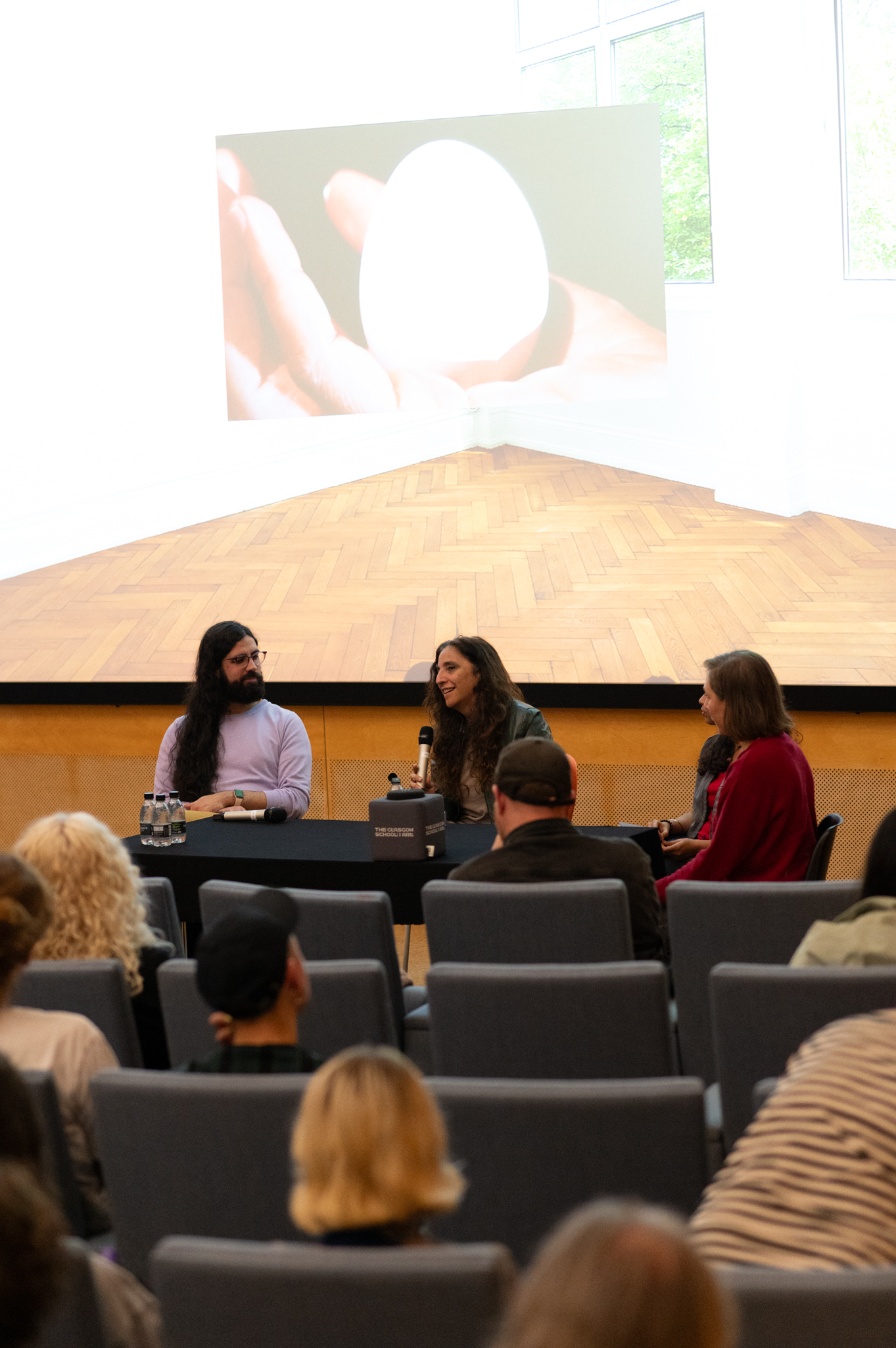 Beatriz Santiago Muñoz talks into a microphone whilst seated at black table in front of an audience. Three other people are also sat at the table. Behind them a projected image of a room with herringbone patterned wooden floor with a superimposed image of an hand holding an egg in its palm