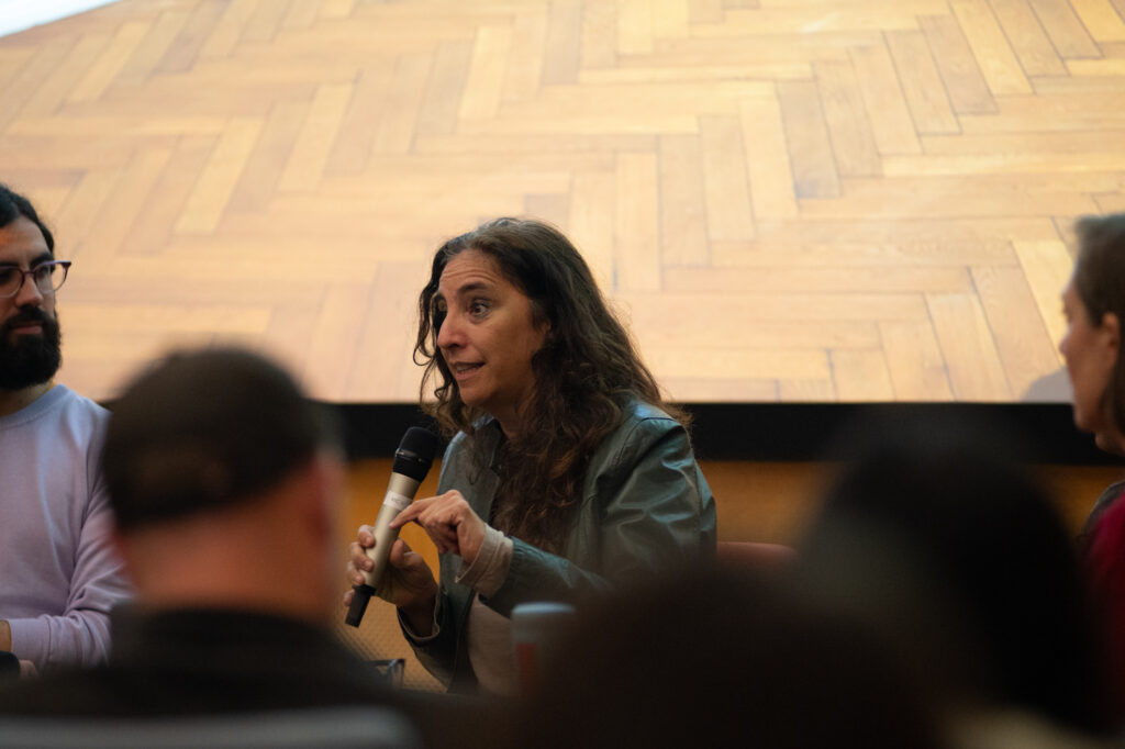 Beatriz Santiago Muñoz talks into a microphone whilst seated in front of an audience. At the back we can see the bottom of a projected image which has a herringbone patterned wooden floor.