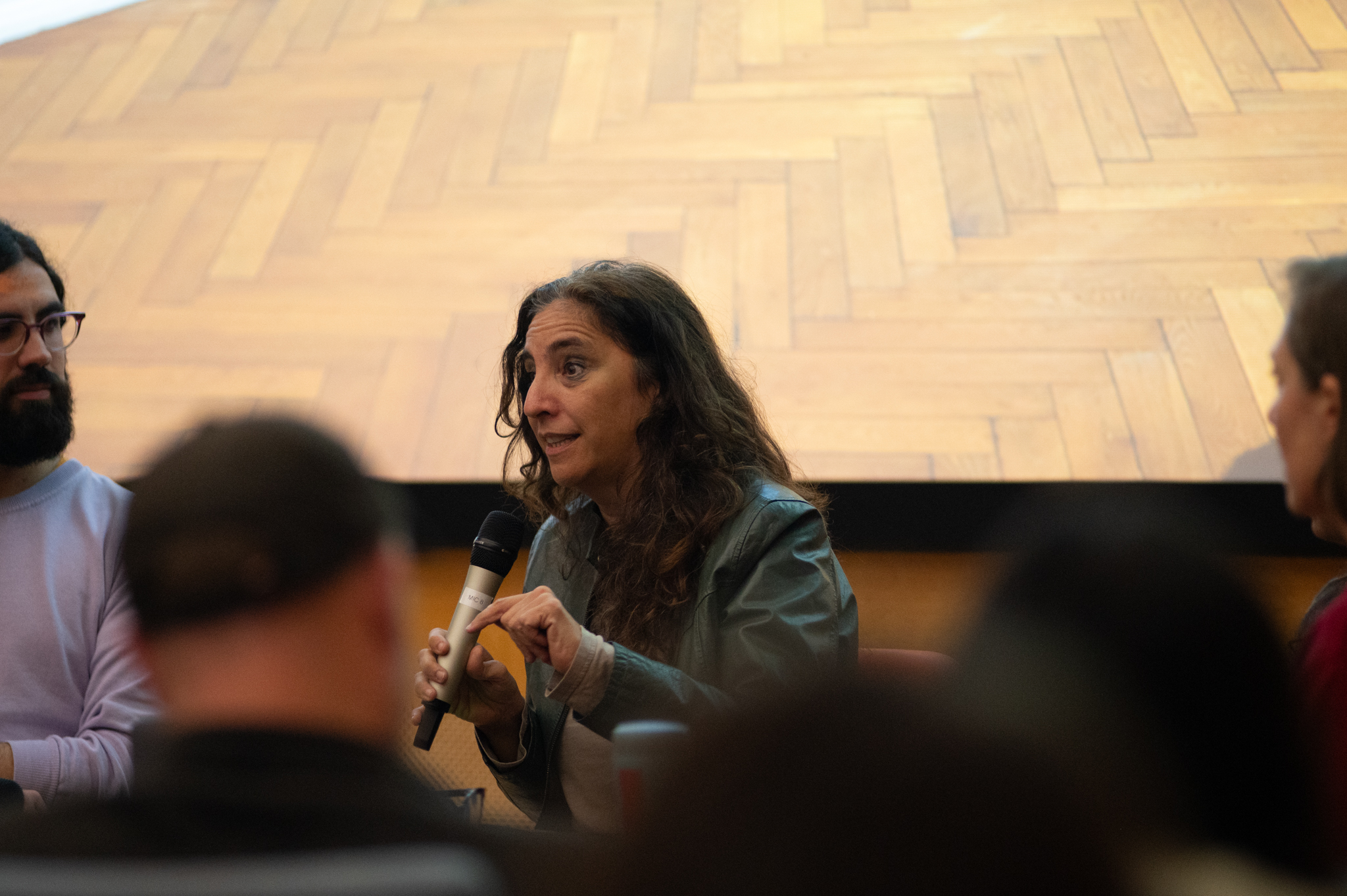 Beatriz Santiago Muñoz talks into a microphone whilst seated in front of an audience. At the back we can see the bottom of a projected image which has a herringbone patterned wooden floor.