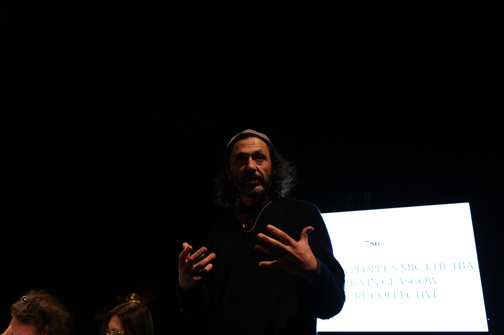 A person stands, gesturing expressively with their hands open in front of them. behind them is a glowing white and faint text