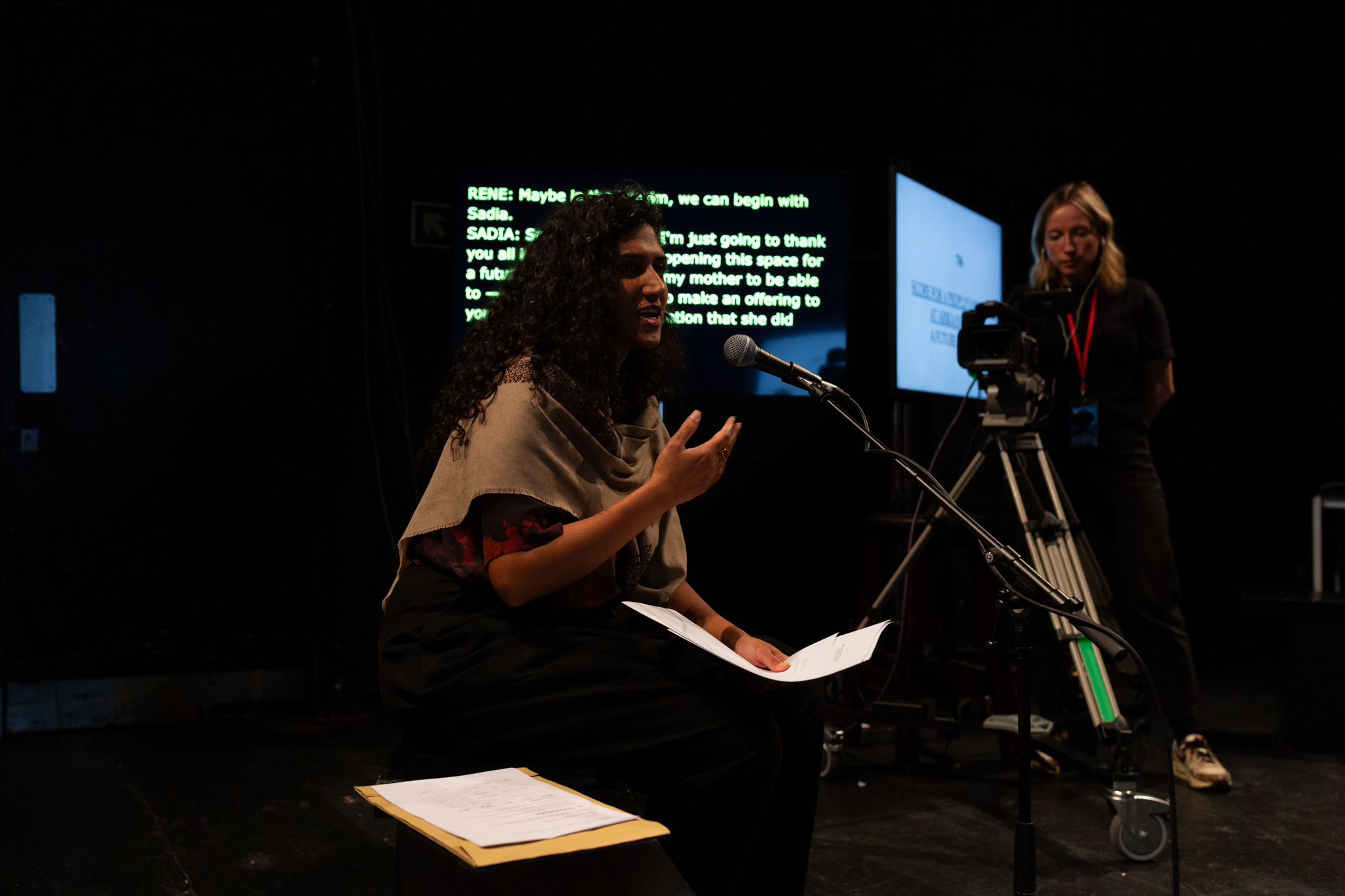 A person is seated talking into a microphone, they are holding a white papers in their hand and an envelope sits next to them. To their left stands a person operating a camera and two screens, one with yellow text and another with a white image & blurred text