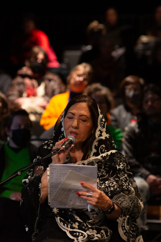 A person sings into a microphone, they are holding a white papers that they are looking at. They wear an ornate black and white scarf that is draped over the back of their head and wrapped loosely around their body.