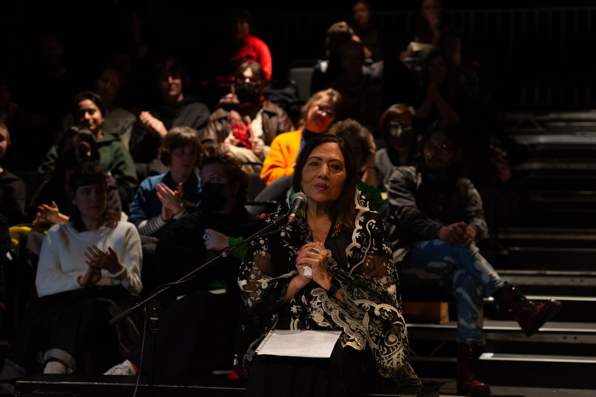 Someone sits infront of microphone with their hands clasped together and a white sheet is sat on their lap. An audience behind them claps.